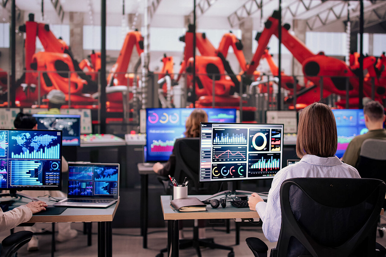 Mitarbeitende an mehreren Bildschirmen mit Dashboards und Diagrammen in einem Kontrollraum, im Hintergrund orangefarbene Industrieroboter.