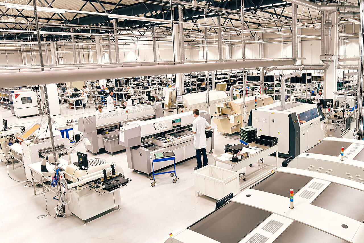 Weitläufige Elektronikfertigungshalle mit mehreren Bestückungsanlagen und Mitarbeitenden in Arbeitskleidung an den Maschinen.
