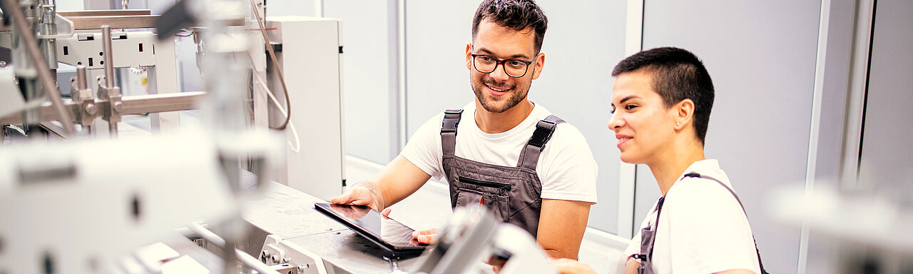 Ein Mitarbeiter mit Brille und Latzhose und eine Kollegin schauen lächelnd auf ein Tablet an einer Montagestation in der Produktion.