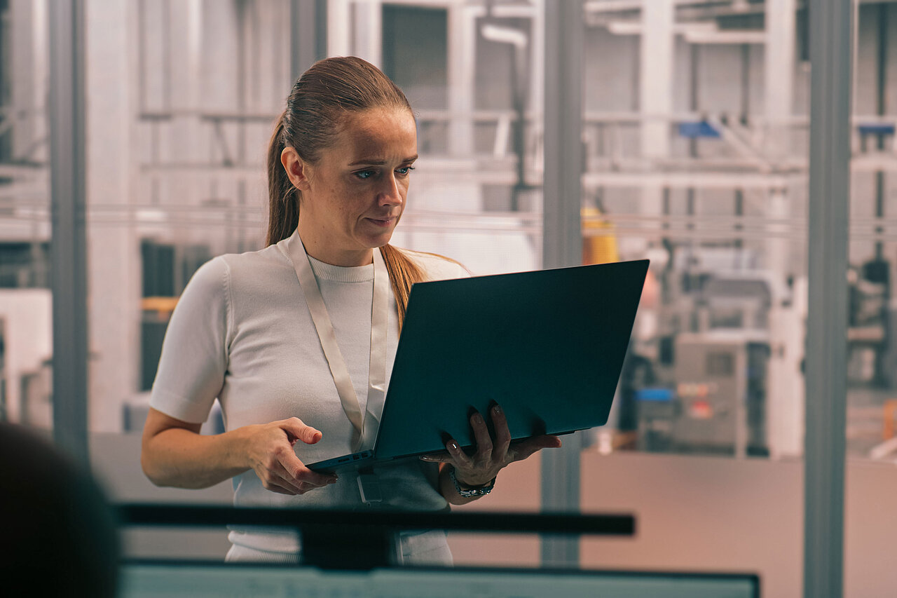 Frau mit Laptop in der Hand steht in einer Produktionsumgebung mit Maschinen und Robotik im Hintergrund.