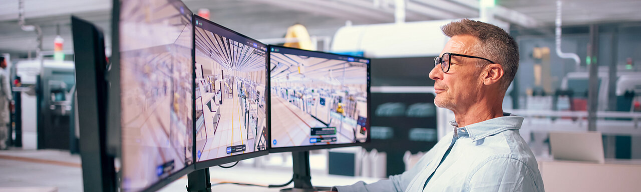 Mann mit Brille sitzt an einem Schreibtisch vor drei Monitoren, auf denen eine digitale 3D-Ansicht einer Produktionshalle zu sehen ist
