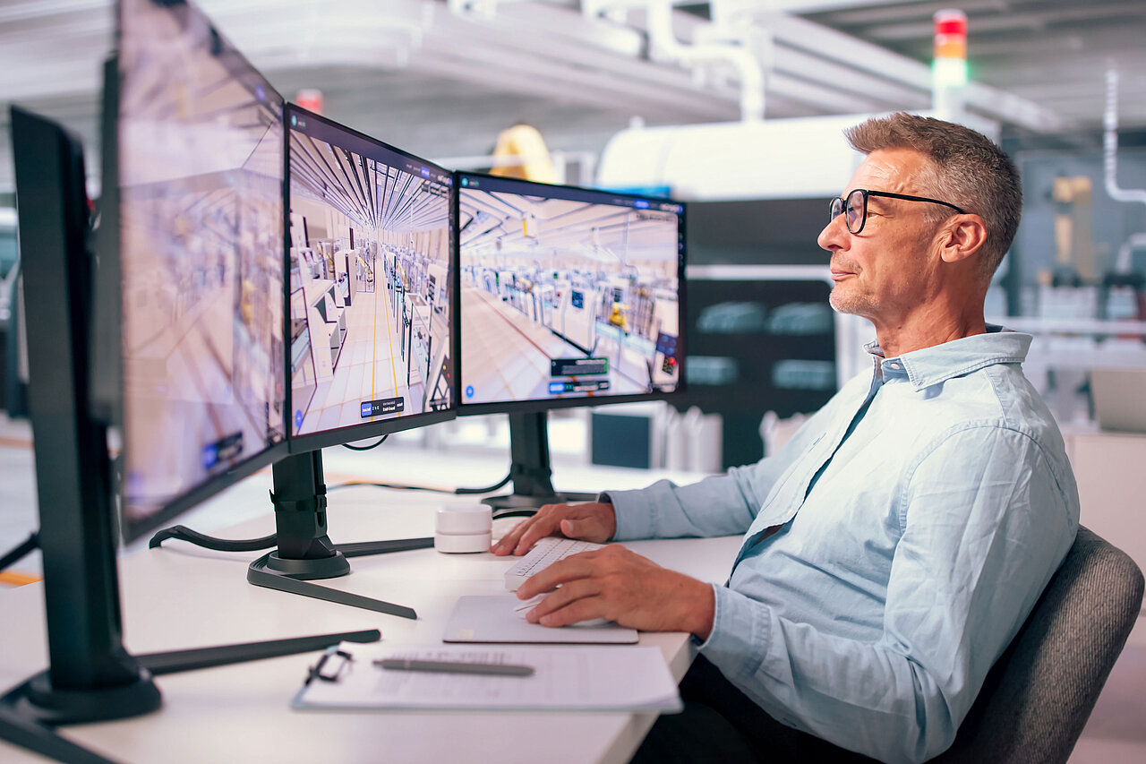 Mann mit Brille sitzt an einem Schreibtisch vor drei Monitoren, auf denen eine digitale 3D-Ansicht einer Produktionshalle zu sehen ist